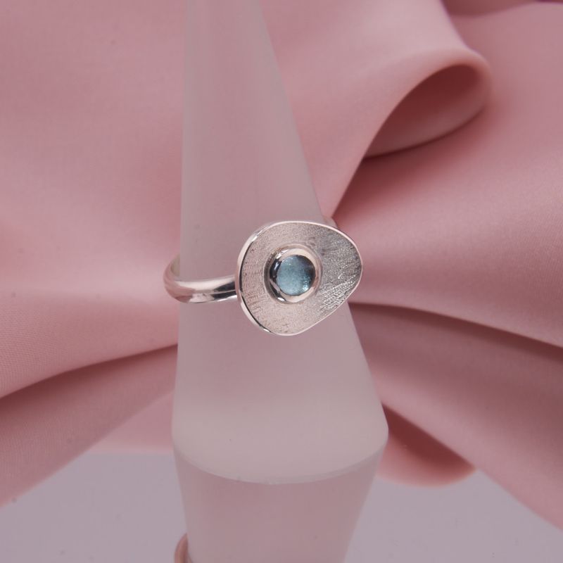 Preview: Präsentation des Rings Isola auf einem Schmuckständer zeigt die skulpturale Form und das Funkeln des blauen Edelsteins.